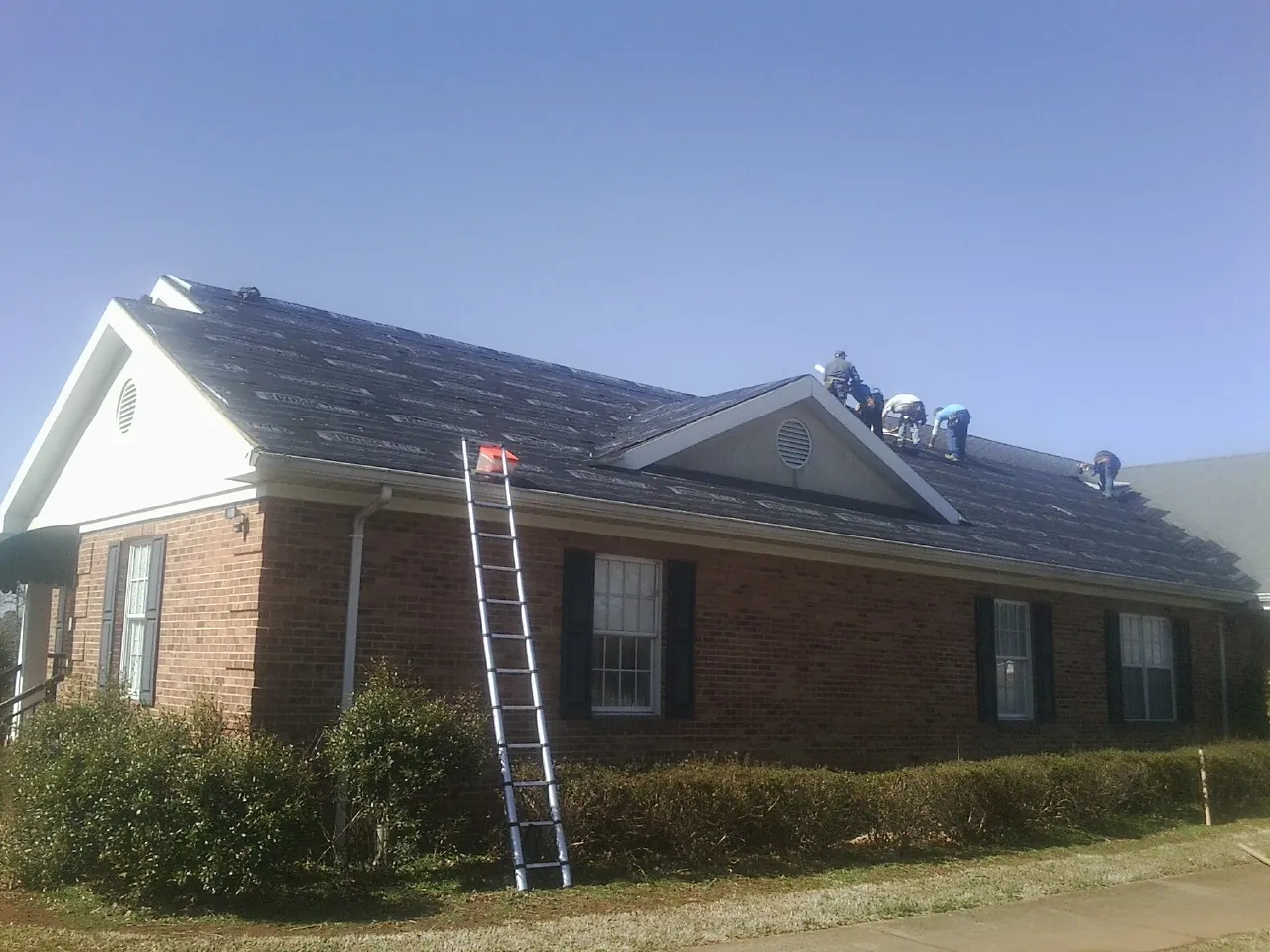 Crew tearing shingles off a brick ranch roof with a ladder set — Metal Roof Repair work by our crew in Van Meter