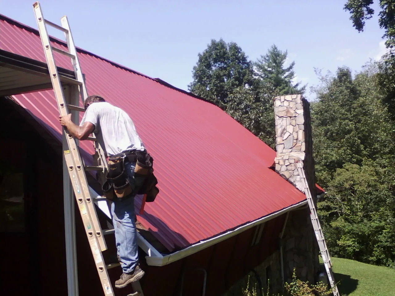 Roofer on a ladder beside a red metal roof and stone chimney — Soffit & Fascia Repair by Roofing Brothers in Van Meter